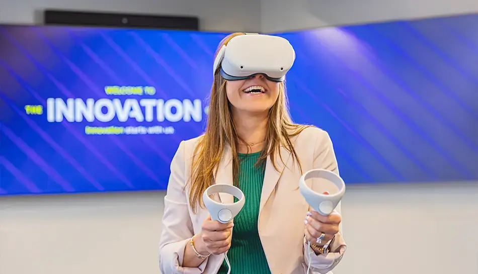 Woman smiling while wearing a virtual reality headset and holding two controllers in front of an innovation-themed display.