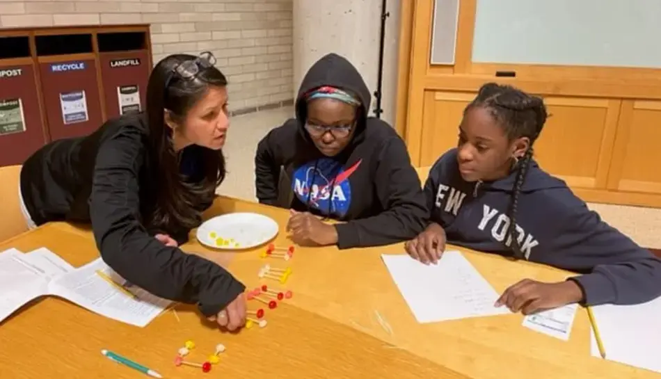A woman guides two students through a hands-on activity using small colorful pieces and wooden sticks at a classroom table.