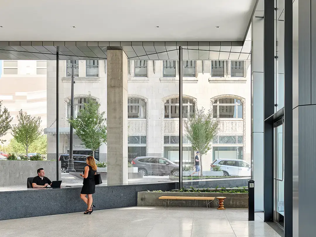 woman standing speaks to man sitting with laptop in a building lobby with view of trees and city street 
