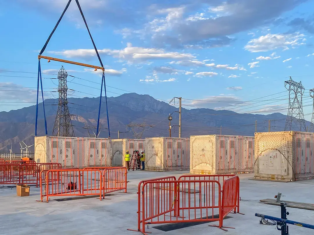 storage unit staging on job site with crane, safety fencing, with lattice transmission towers and mountains in background