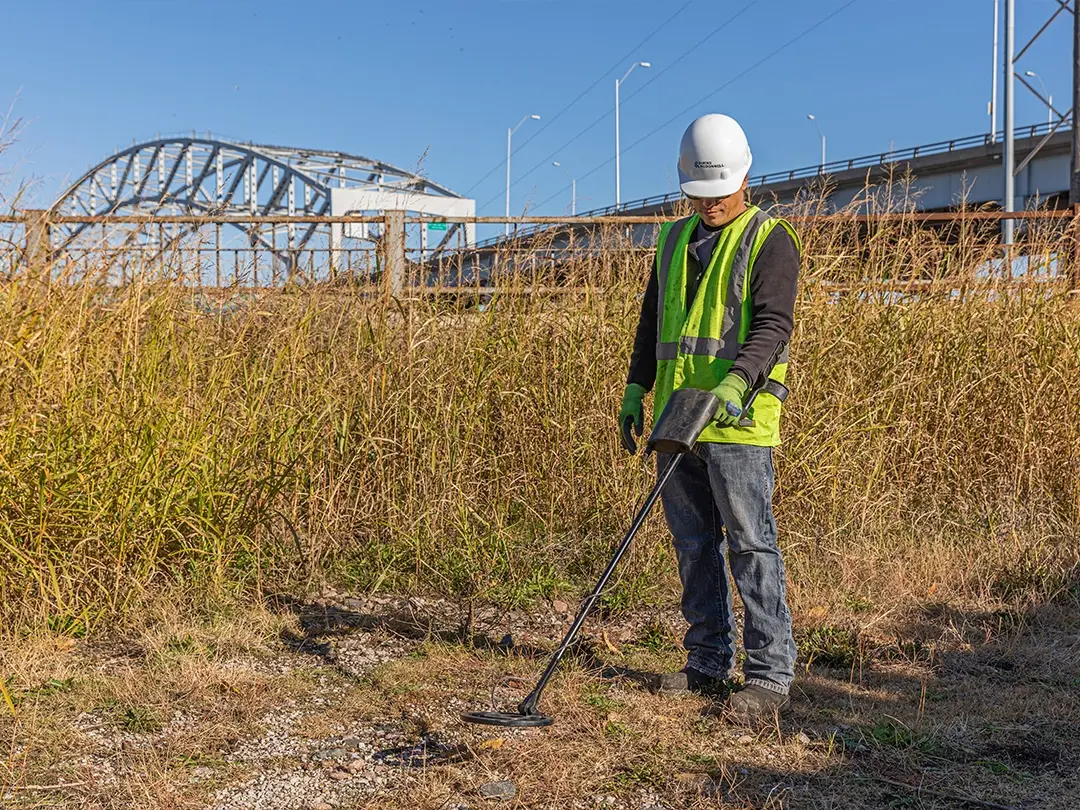 worker using metal detector in grassy area with bridge in background