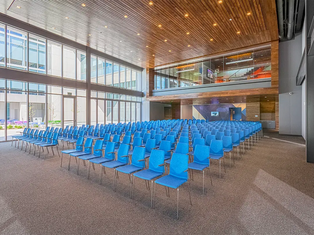 chairs in place in an auditorium with wood ceiling and glass conference room windows overlooking space; exterior windows look out on sunny common space with tables and chairs