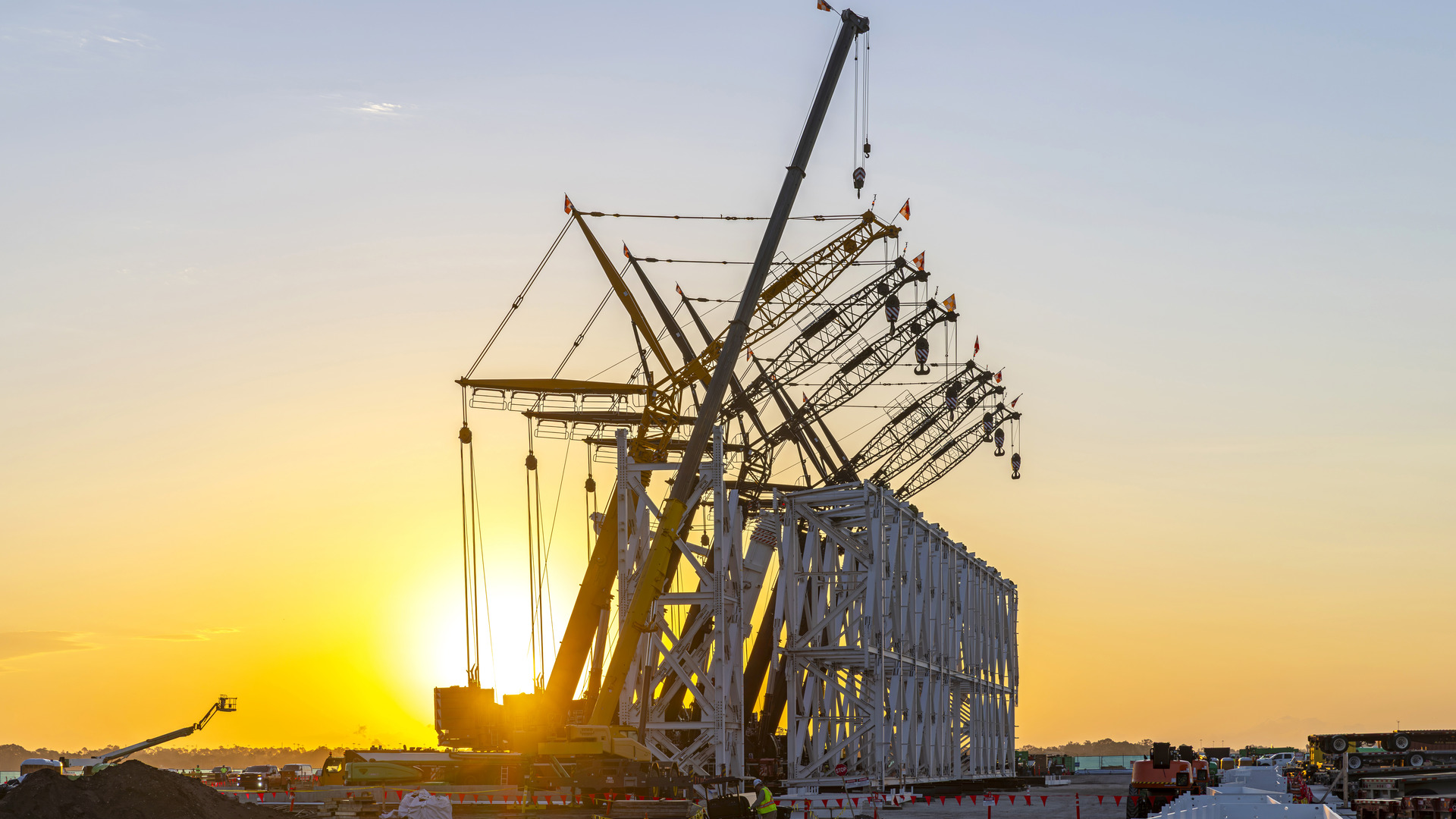 Multiple cranes stand beside a large steel framework at an airport construction site, silhouetted against a golden sunset.