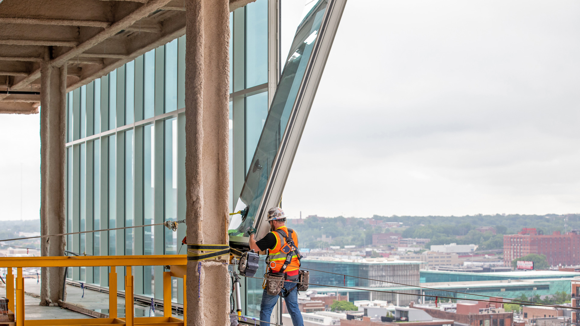 Construction worker installs a large glass facade panel on a high-rise building, secured by safety equipment with a cityscape visible below.