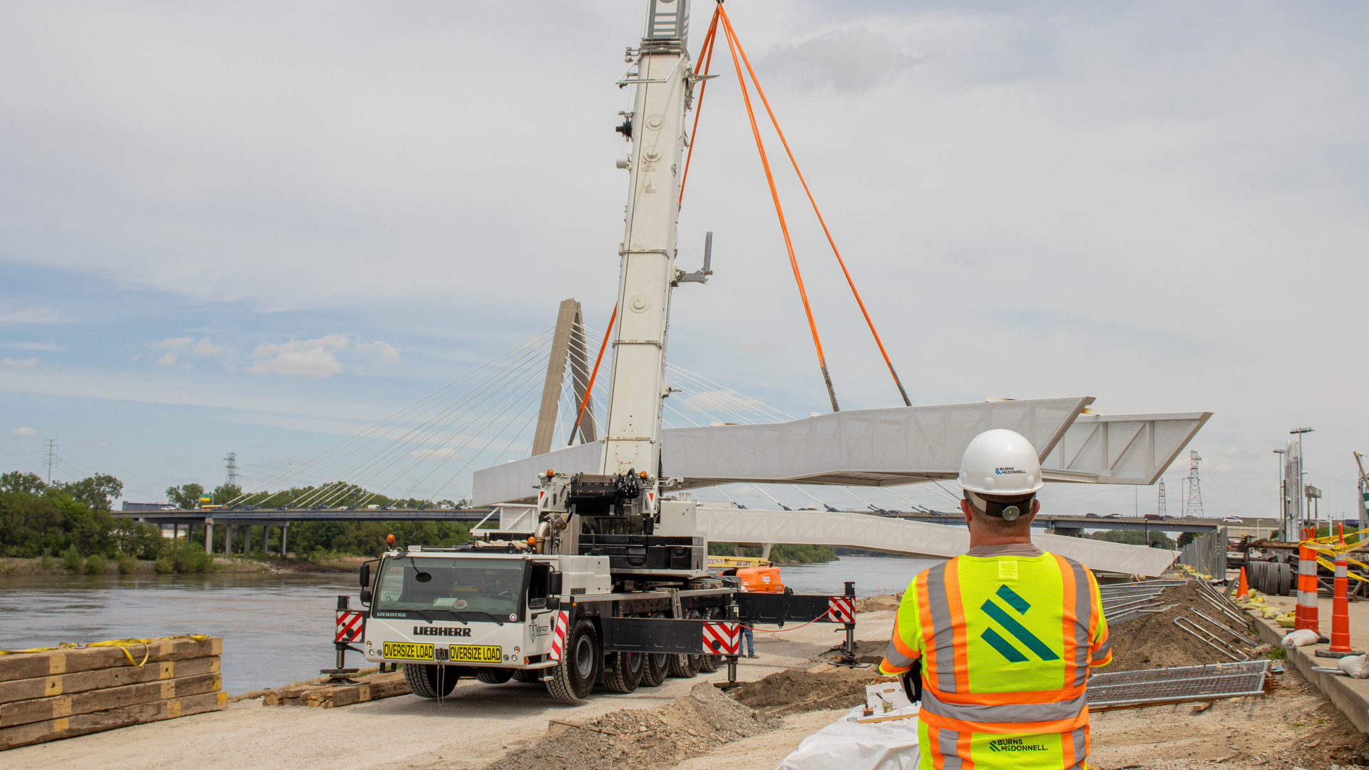 Construction worker oversees a crane lifting a large steel bridge section beside a river.