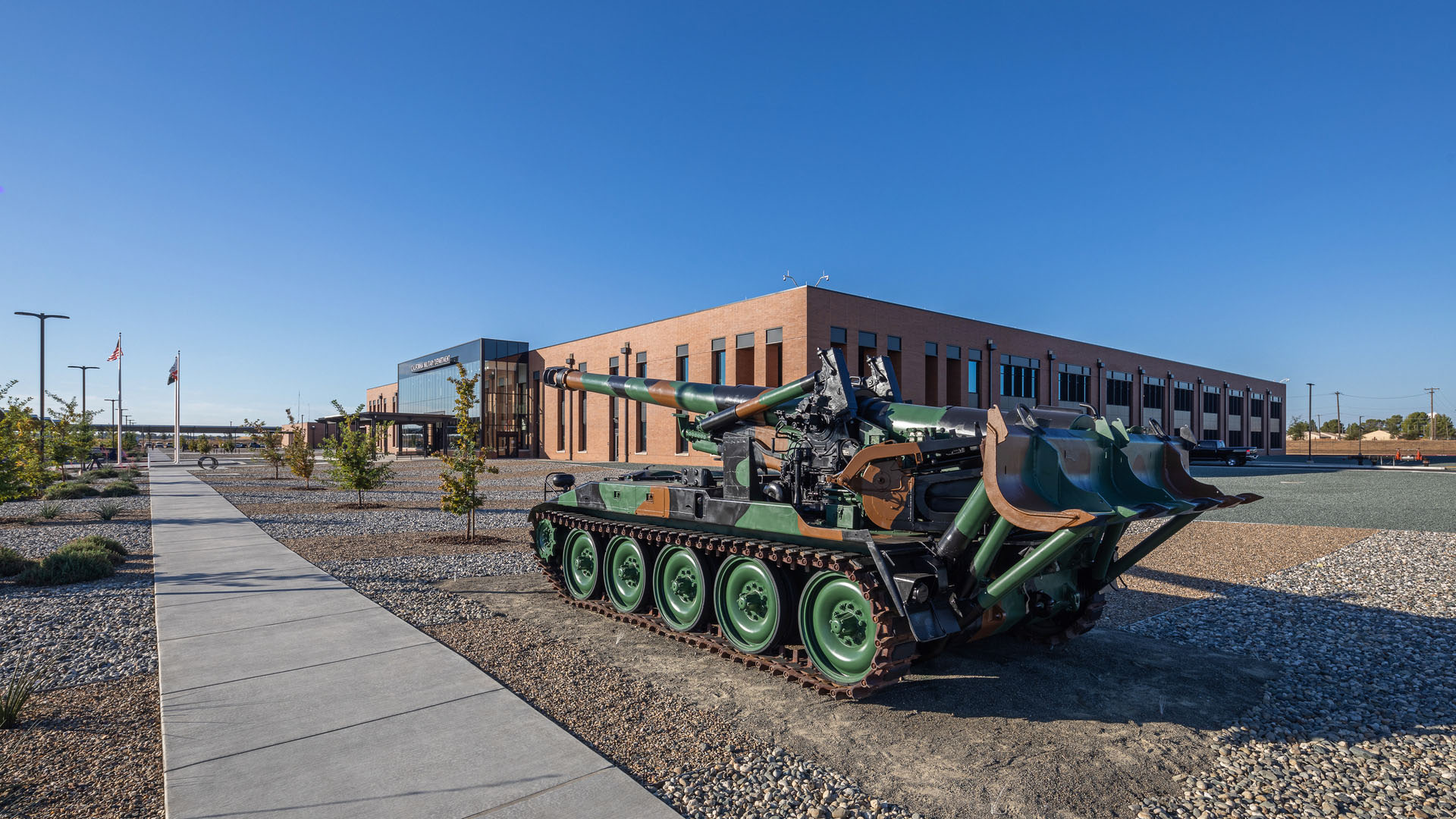 Tracked military artillery vehicle displayed outside a modern brick facility.