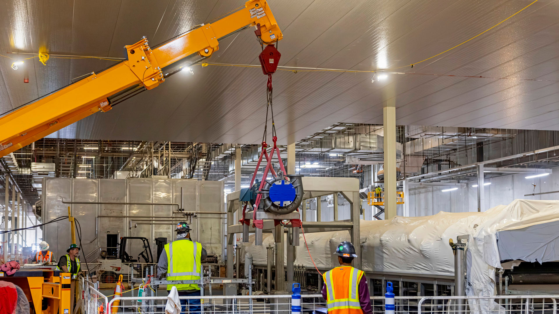 Workers oversee a crane lifting industrial equipment into place inside a large manufacturing facility under construction.