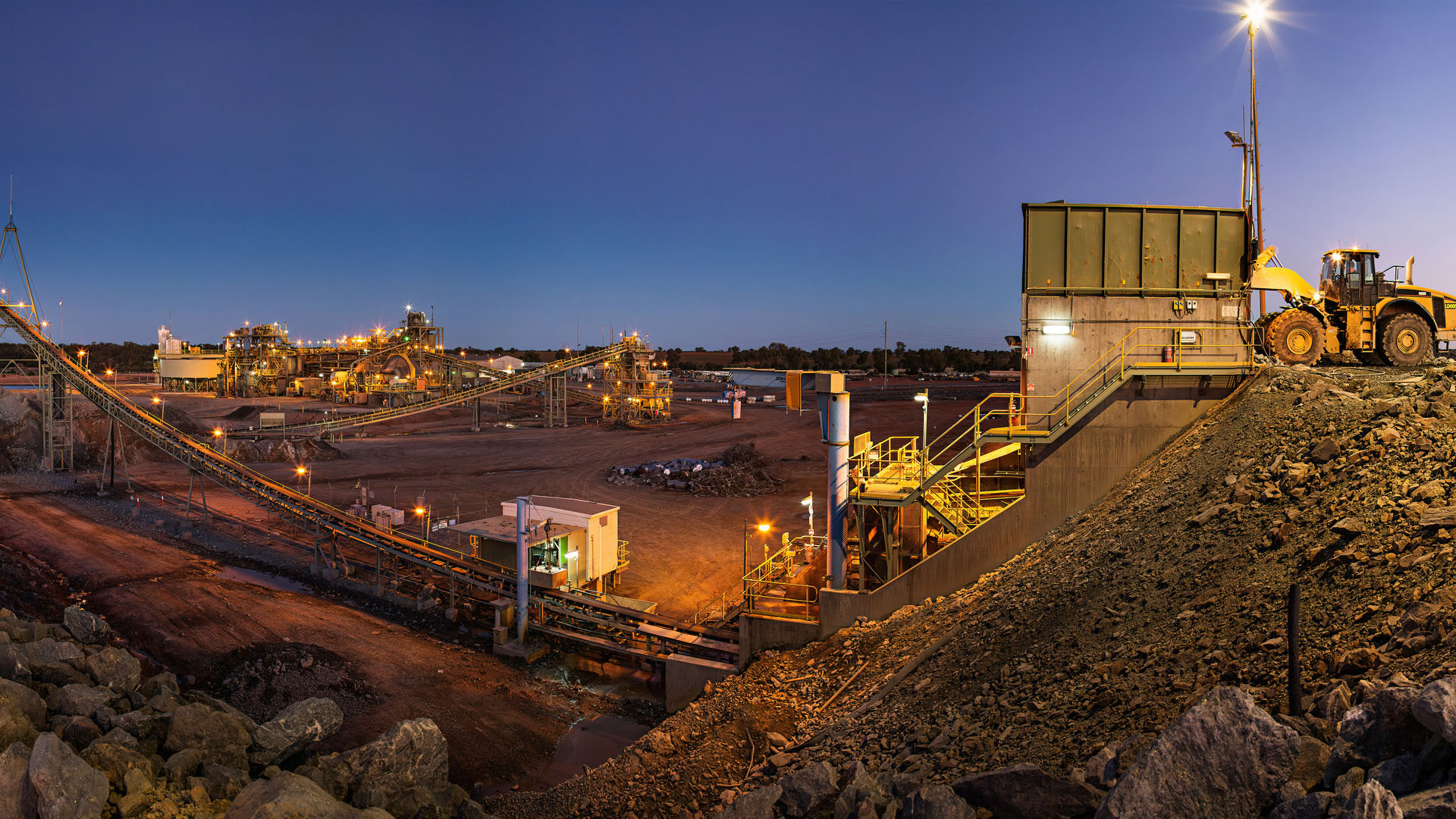 Illuminated mining and mineral processing facility at dusk, with conveyor systems, industrial equipment and a large loader on a rocky slope.