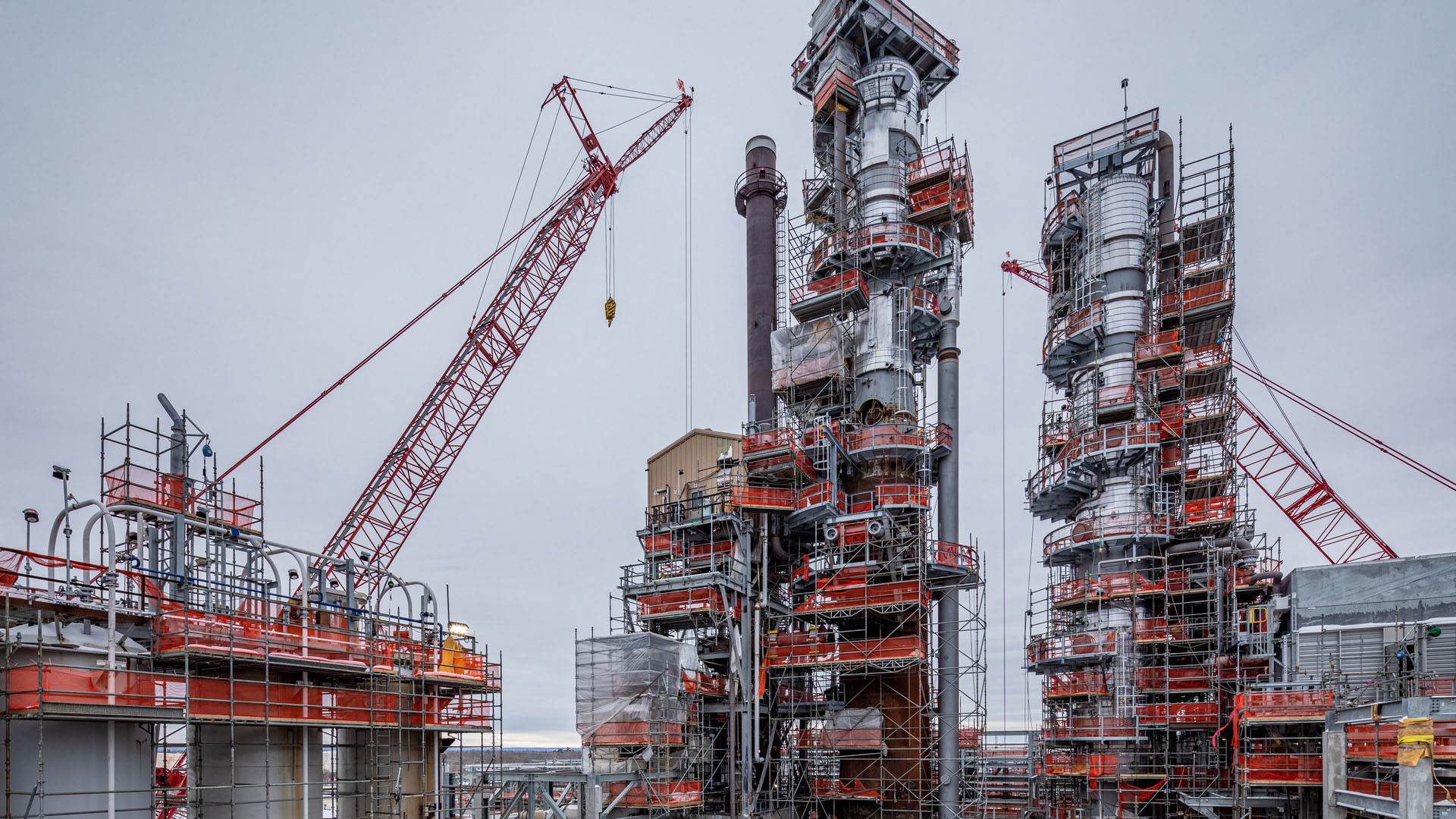Large industrial processing towers wrapped in scaffolding and orange safety barriers stand between red construction cranes.