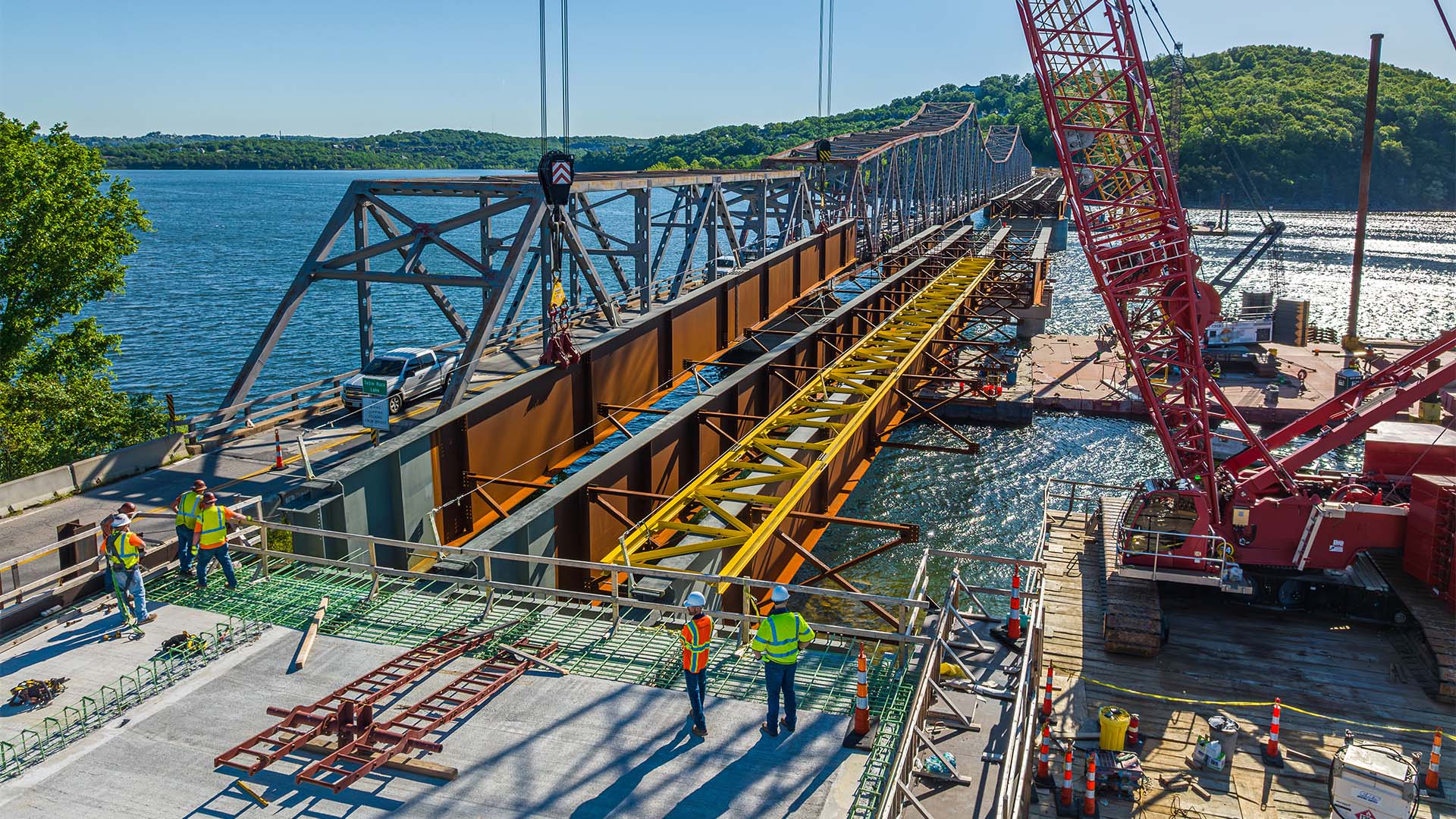 Worker welds steel components at a bridge construction site beside the water.